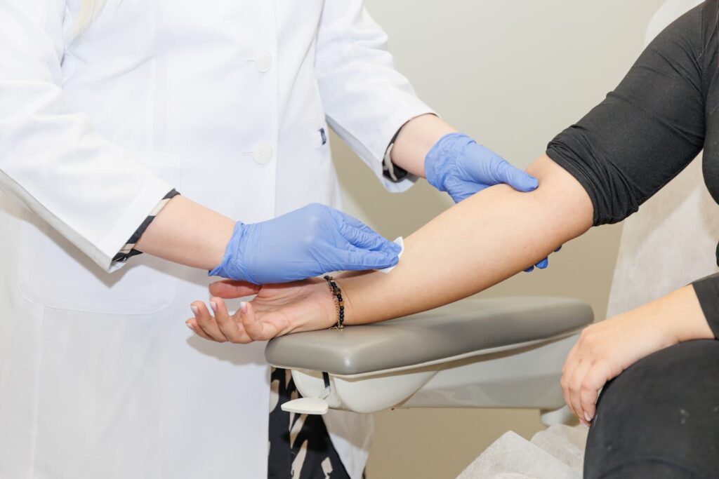 A healthcare professional wearing a white lab coat and blue nitrile gloves uses an alcohol wipe to sanitize a patient's arm. The patient's arm is resting on a grey medical armrest in a clinical setting.