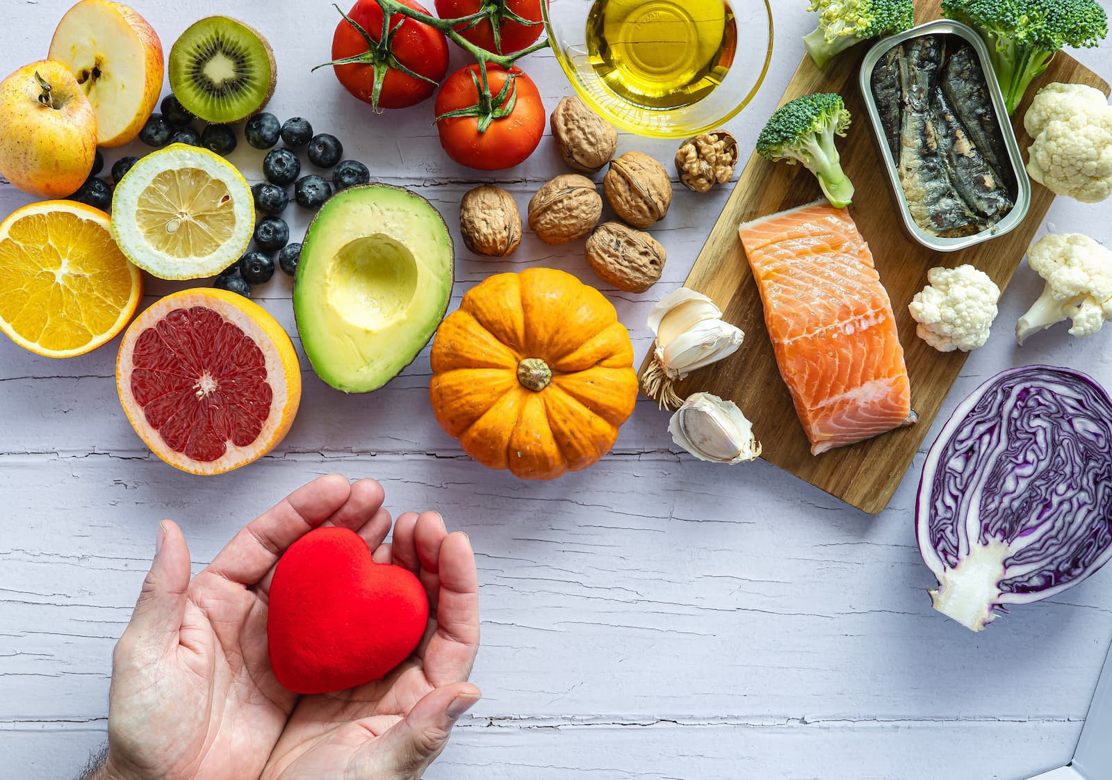A top-down flat lay of various nutrient-dense foods on a light wooden surface. The arrangement includes citrus fruits, berries, avocado, tomatoes, walnuts, a small pumpkin, salmon, sardines, broccoli, and red cabbage. In the bottom left corner, a pair of hands gently cups a small red heart.